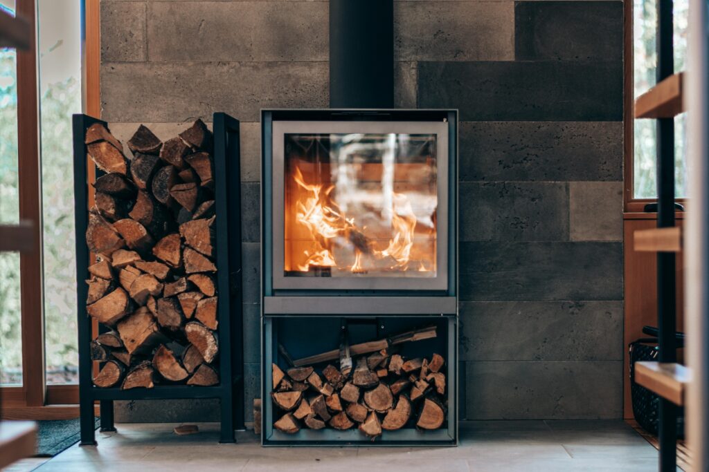 Kamin in dem Holz brennt mit einem Holzstapel daneben vor einer Wohnzimmerwand aus dunklen, großen Fließen